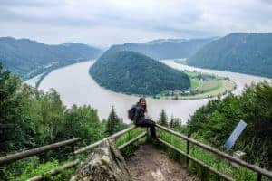 A woman sits on a rail at a viewpoint overlooking the Schlögener Schlinge bend in the Danube River in Austria.