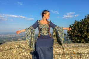 A woman, dressed in a navy blue medieval dress with green-golden decorated bodice and sleeves, sits on a golden rock wall that is elevated above a valley, seen in the background.