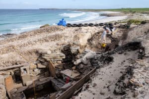 An archaeologist carries a yellow bucket about a hollow ditch at an excavation site on the light sandy coastline of the Orkney Islands in Scotland.