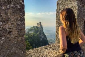 A woman looks through an opening of a stone wall out towards a castle on a hilltop in San Marino.