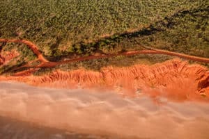 An arial view of the light and bright orange sands of the Outback coastline in Western Australia. The desert sands merges with the green forest that backs it.