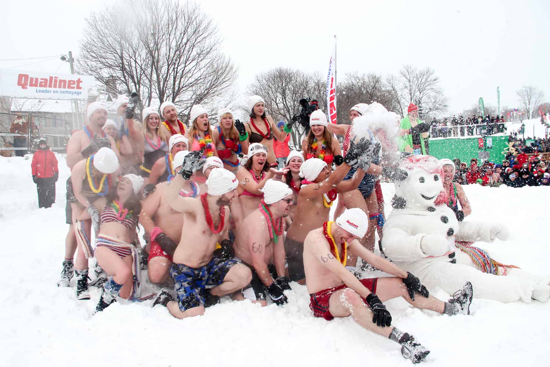A group of people in swimwear and white hats sit in the snow with the snowman figure, Bonhomme, during the Snow Bath at the The Carnival de Quebec.