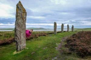 A woman in a pink jacket peeks out from behind a tall, rectangular rock on a grassy mound – one of five stones seen in this picture that belong to the Ring of Brodgar stone circle in the Orkney islands, Scotland.