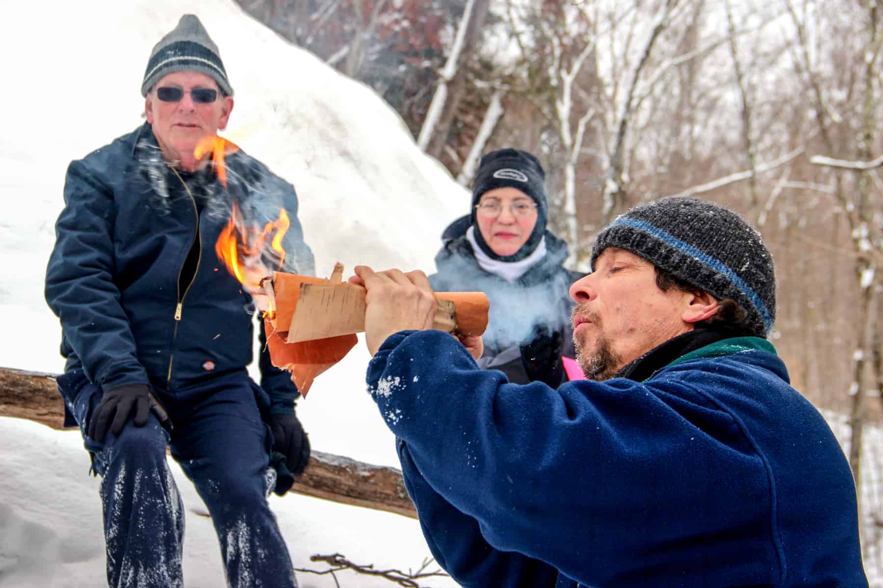 Two people look on as a man blows into a roll of paper to demonstrate how to start a fire in the snow.