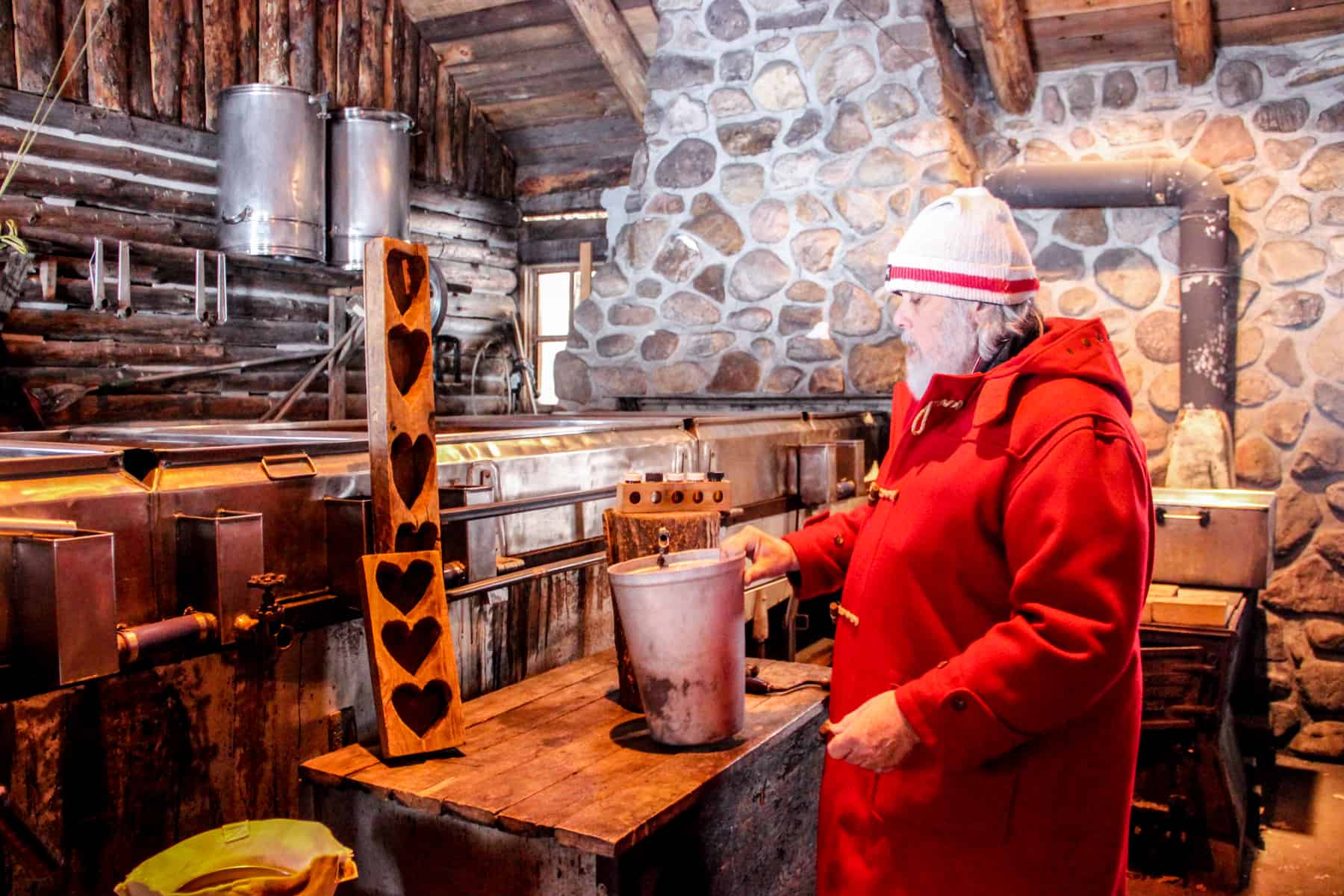 The owner of the Sucrerie de la Montagne sugar shack, dressed in a red coat and white hat, stands within a wood and stone storage building holding a metal bucket on maple syrup. 