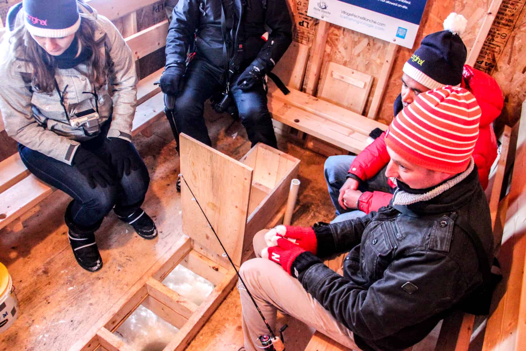 Four people sitting in a wooden hut in front of a row of holes drilled into thick ice. One man has a fishing rod next to him in preparation for ice fishing. 