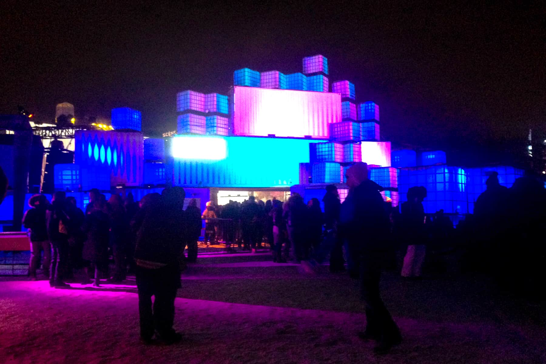 People walking towards a building made of blocks of blue and purple neon light – a night scene from the Igloofest in Montreal.