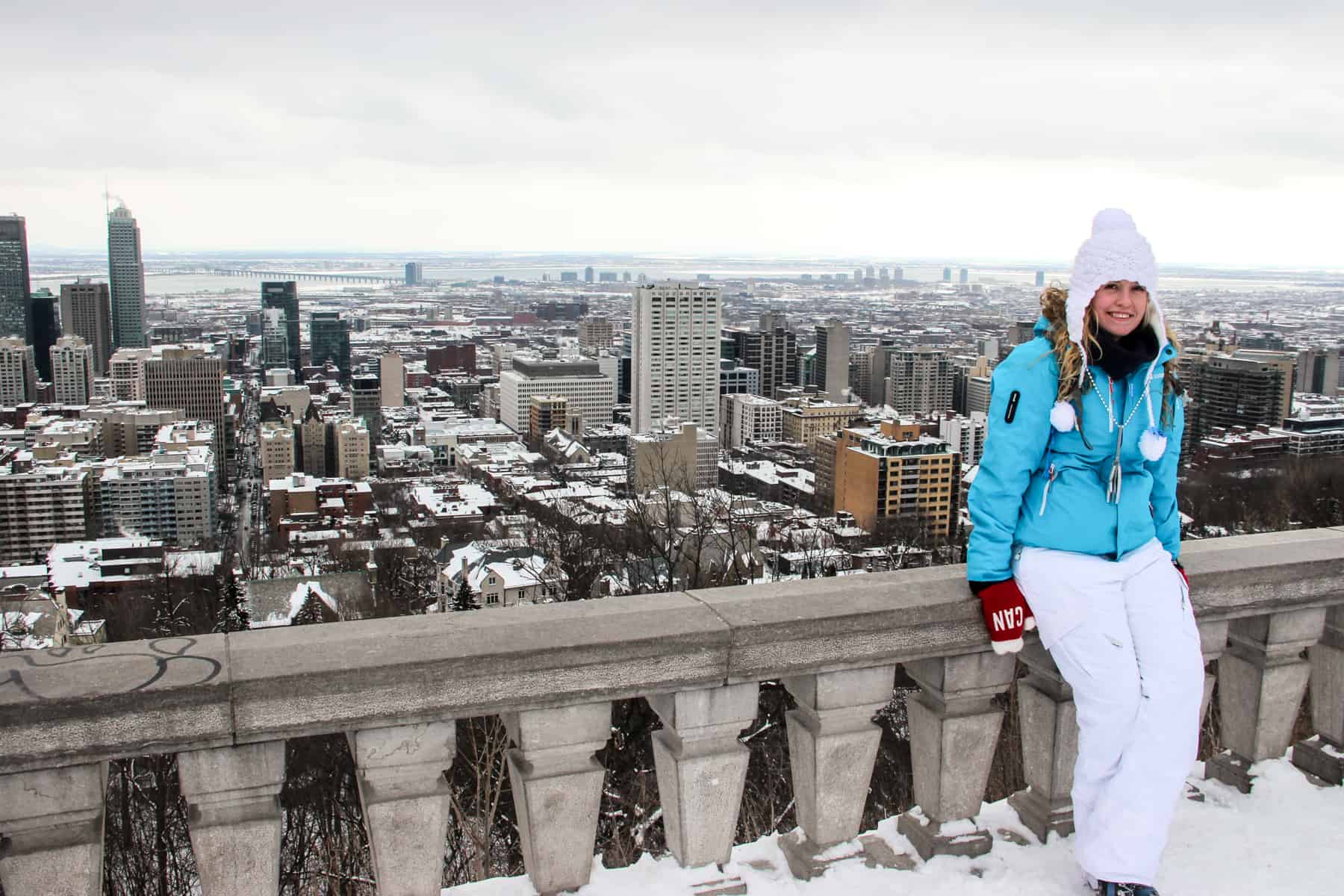 A woman in blue and white ski clothing leans on a stone wall with Montreal city in the background. 