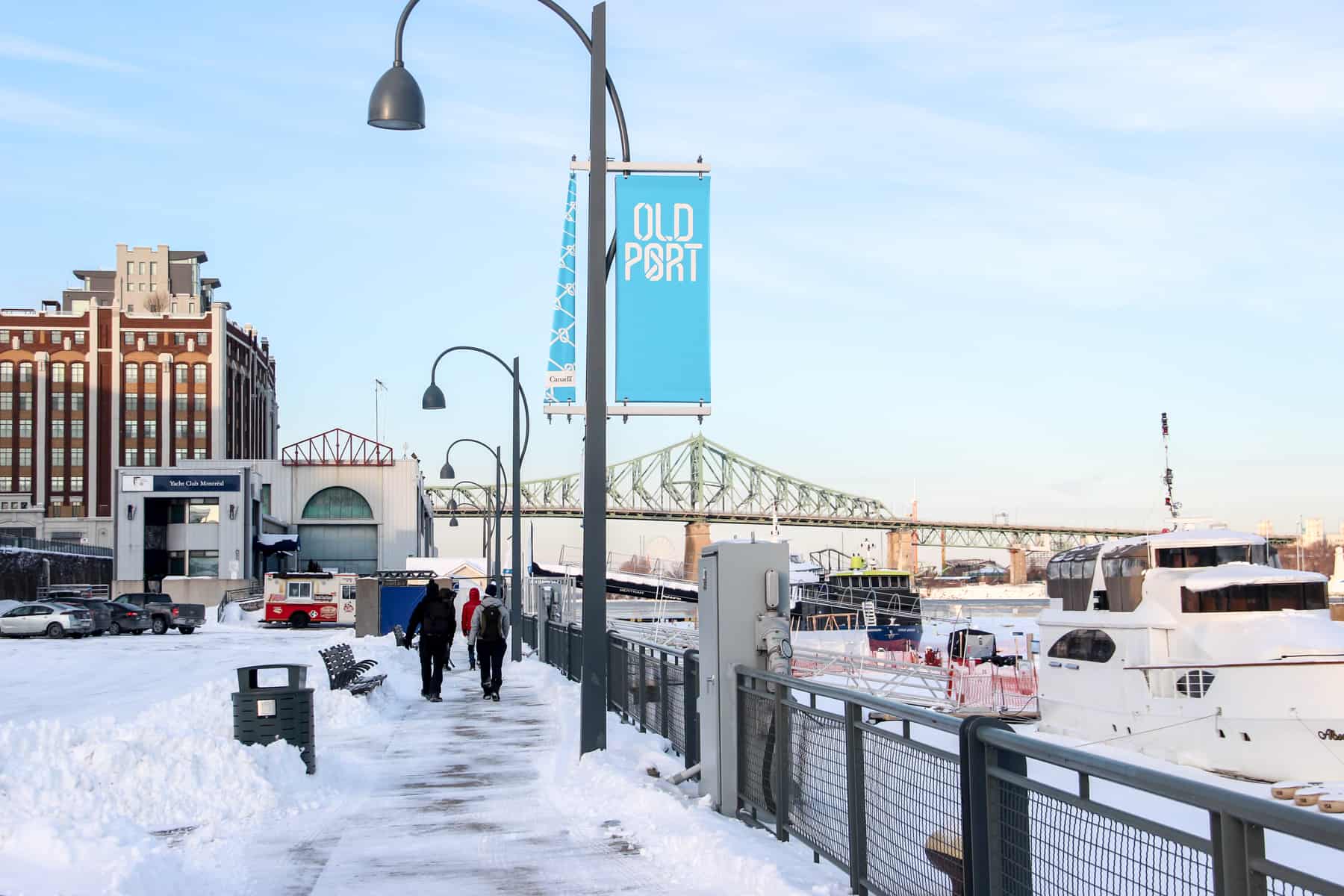 People walking on the snow covered pathway next to the frozen Old Port of Montreal, where boats rest in the ice. 