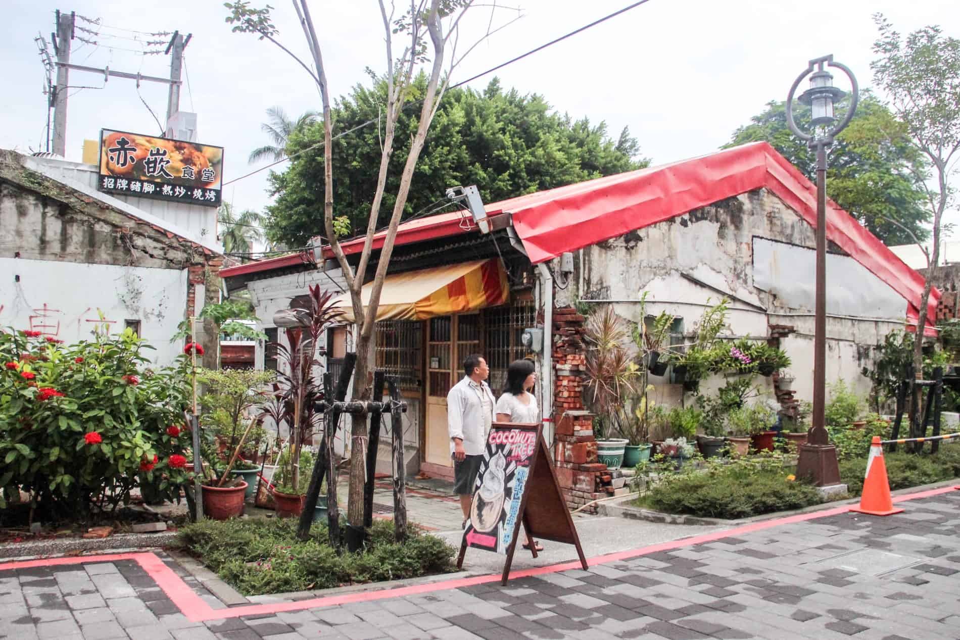 A couple walk past on an avenue past an old house with a red fabric roof in Tainan cirty. 