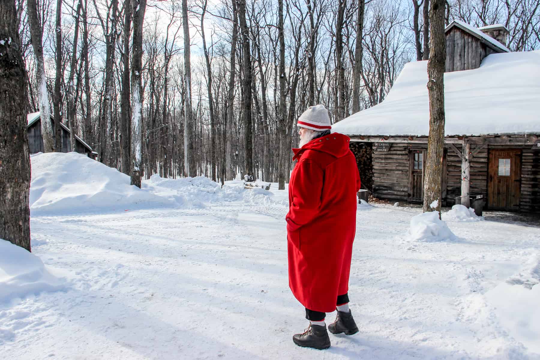 A Santa Claus like figure in a red coat and white hat walks past a wooden hut in a snow covered forest. 