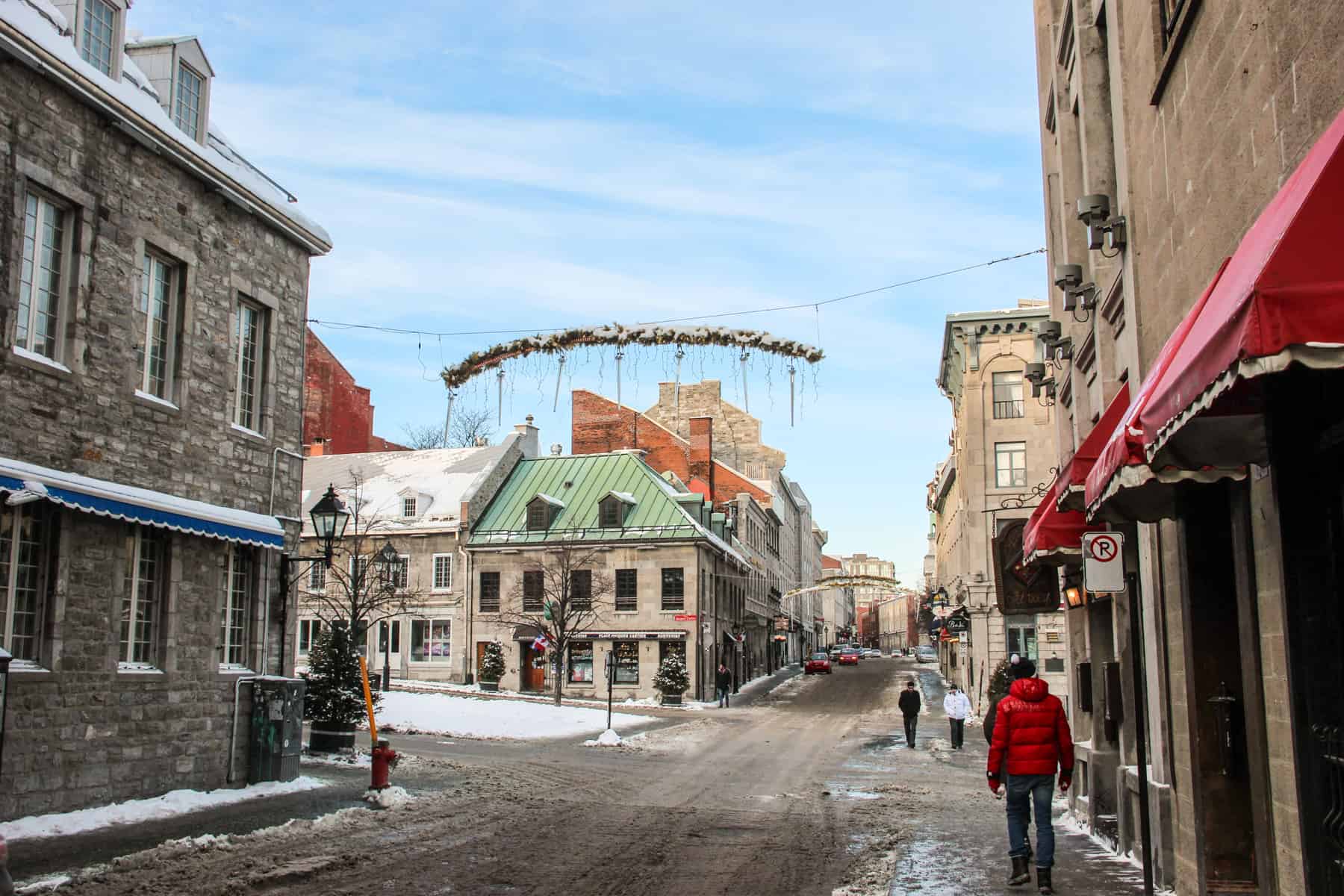A person in a red jacket walks on the side of the street within a grid of golden brown stone buildings in Old Montreal in winter. Christmas decorations hang overhead. 