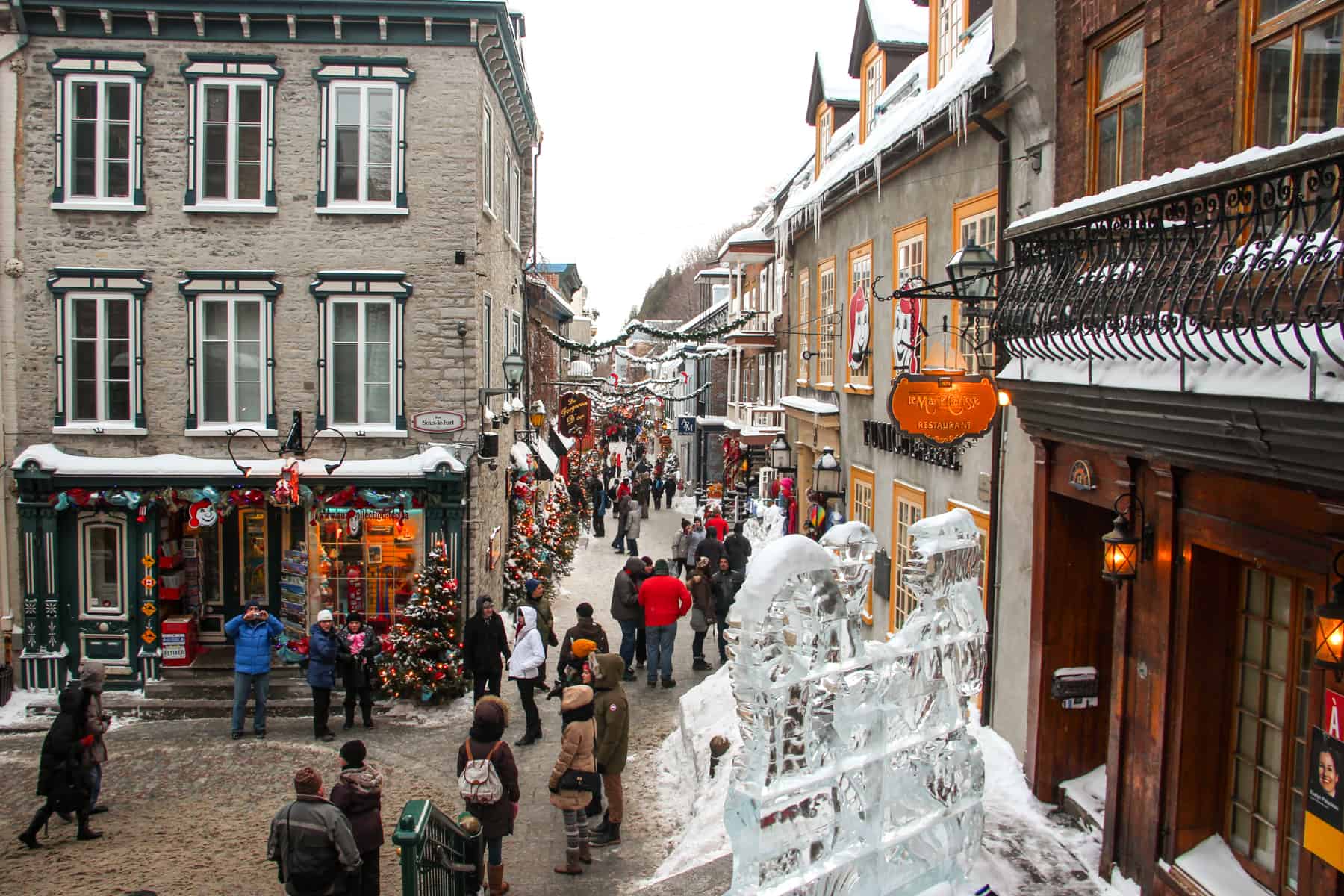 People walking on the streets of old Quebec city in winter, where balconies are covered in snow and an ice sculpture stands in front of a store. 