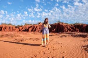 A woman in a maxi dress walking through the striking, ochre orange desert sands in Western Australia, on a landscape backed by rugged red rocks and a bright blue sky.