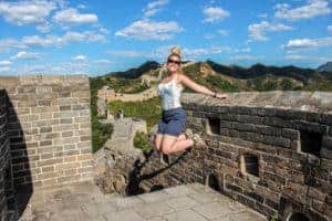 A woman jumps in the air standing on one of the stone turrets of the Great Wall of China. In the distance is the vast green forest.