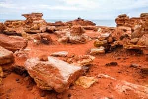 A bundle of bright orange rocks and sand in Brome, Western Australia. In the distance a couple can be seen sitting upon one of the rock looks out towards the ocean ahead.