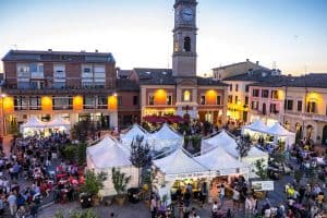 A mass of people gather in a huge square around the white food tents at the Festa Artusiana food festival in Forlimpopoli, Italy. Behind the tents the building lights are a golden orange and the tall column is a clocktower that pokes through the sky.