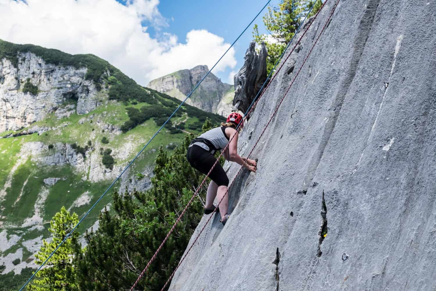 A woman rock climbing on a steep silver flat incline to a backdrop of forest covered mountains.