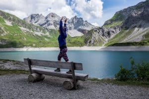 A woman stands, hands raised, on a bench next to the mountain top Lake Lunersee in Austria