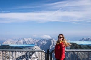 A woman in a red jacket stands at a viewing platform on the Zugspitz mountain, her back to a panorama of mountain peaks.