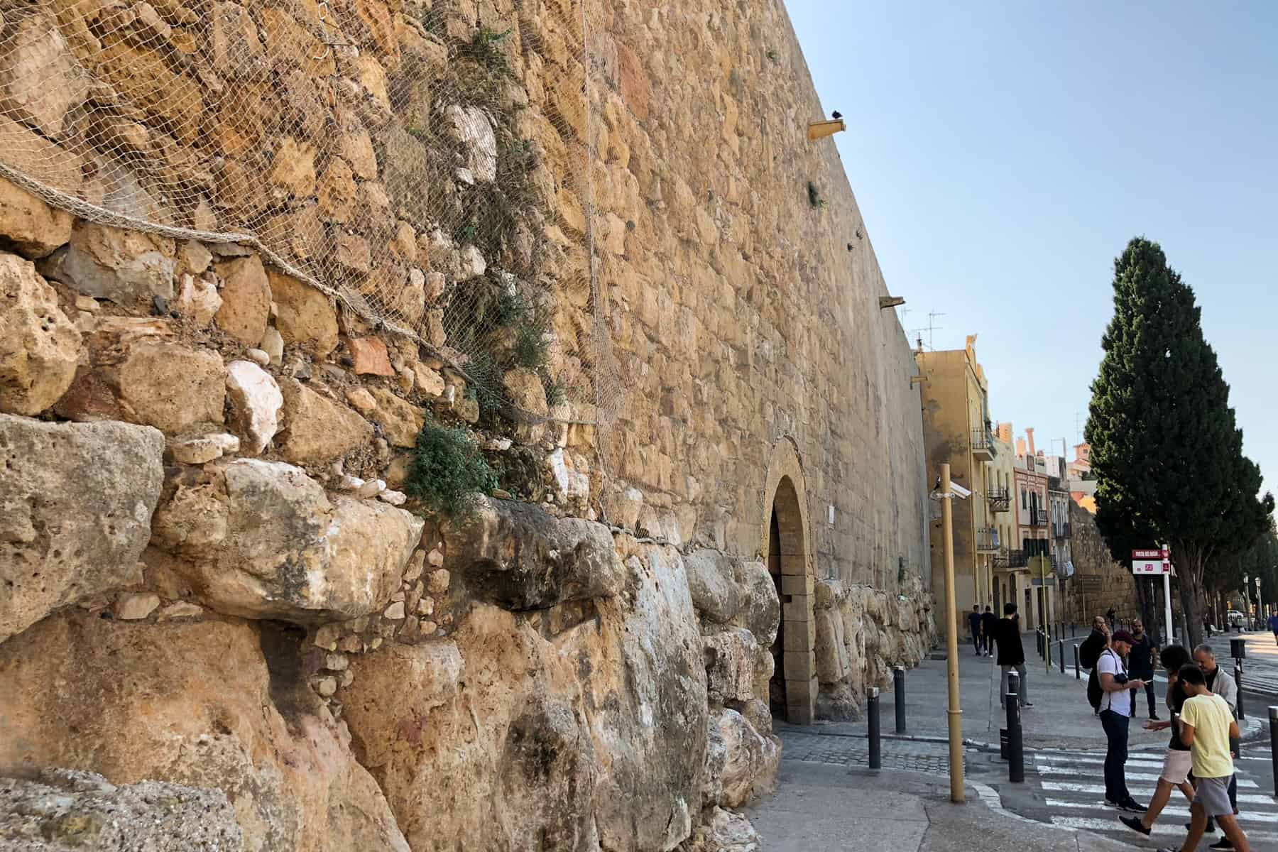 People walk towards a tall wall of Golden stone - the old Roman city walls still visible in Tarragona, Spain