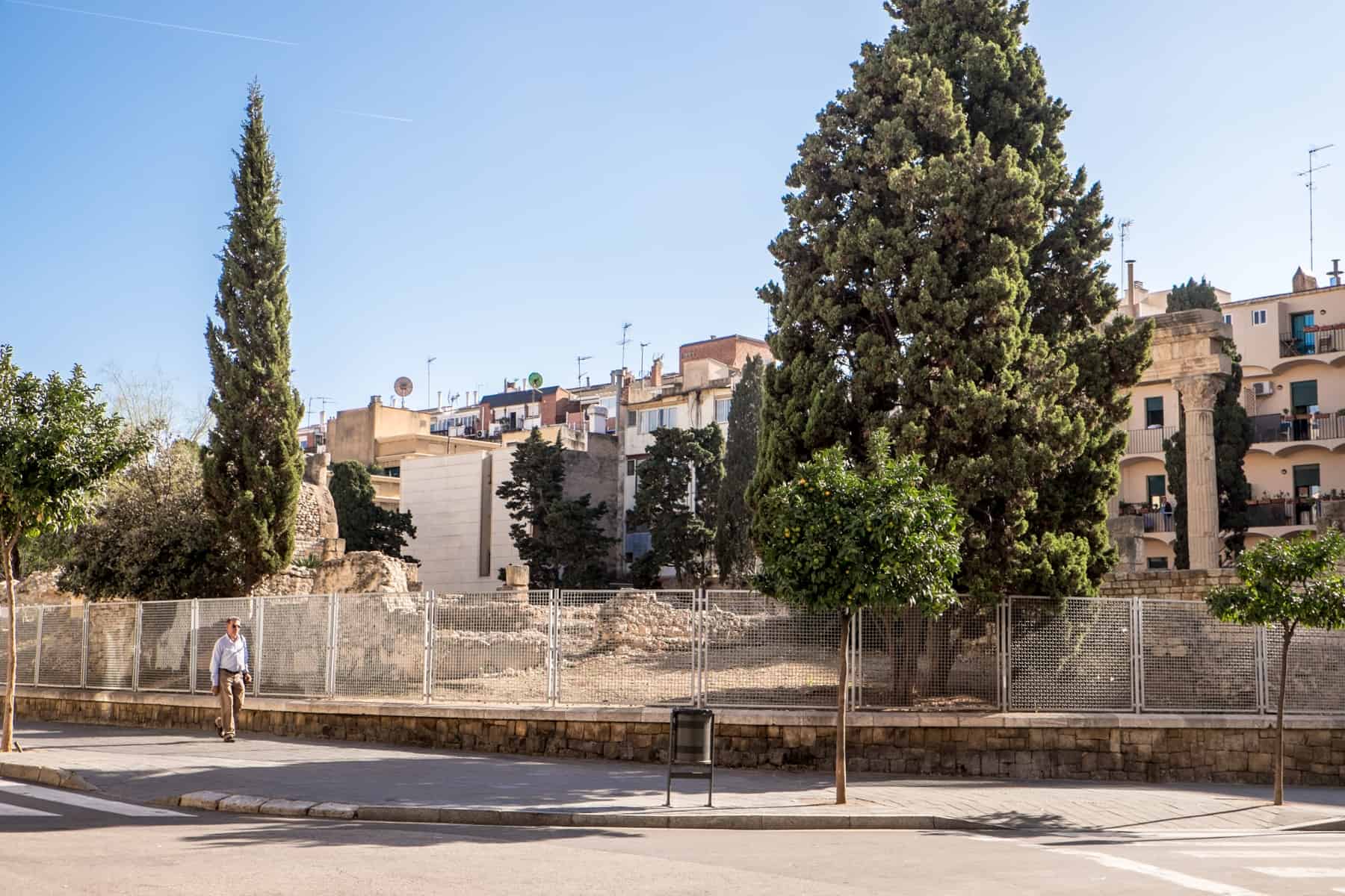 A man in a white shirt walks past a parkland of trees filled with the roman ruins of a square and temple column in Tarragona, Spain