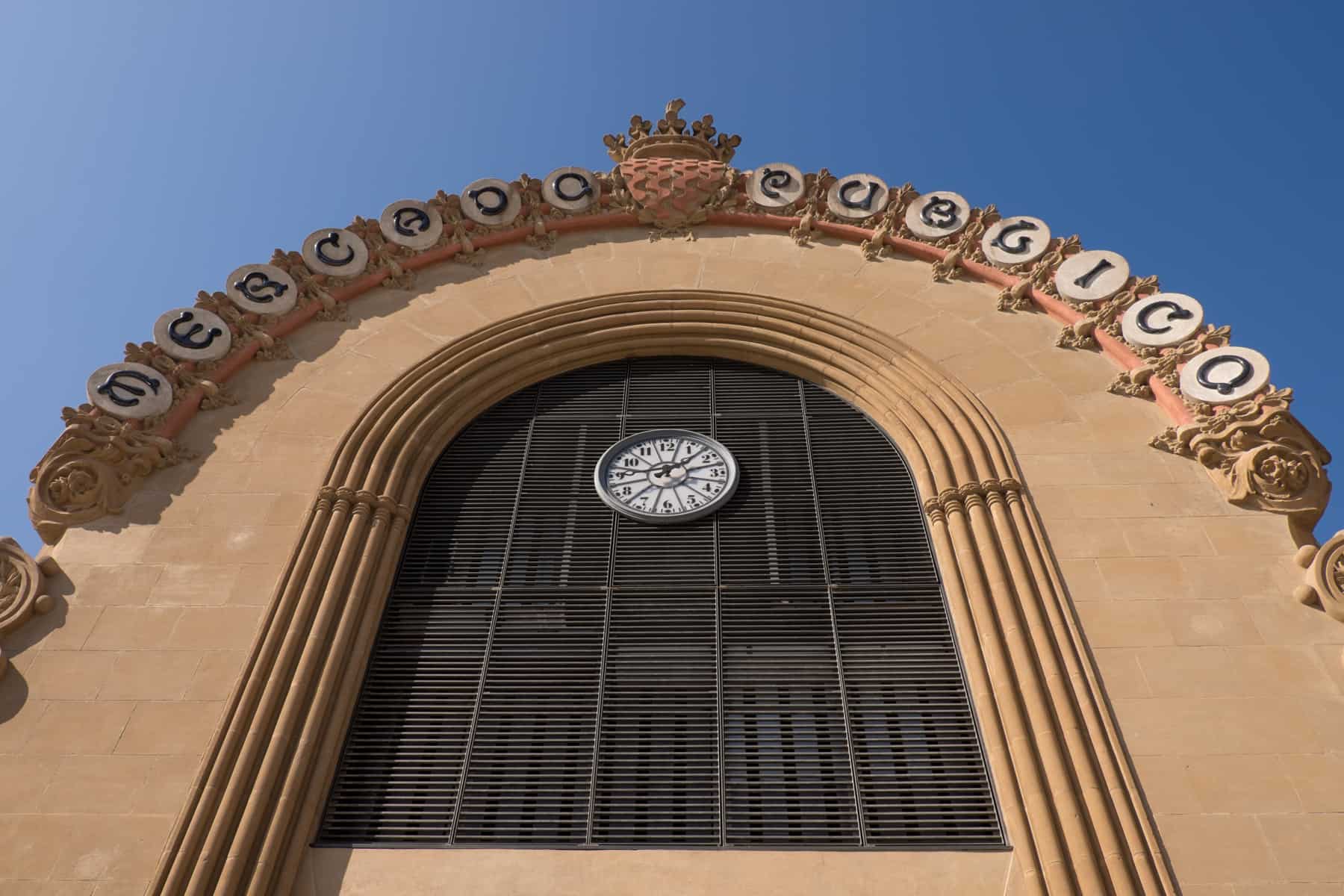 A rounded archway exterior design, with a white clock and round white lettering signage of the Mercado Central de Tarragona Spain 