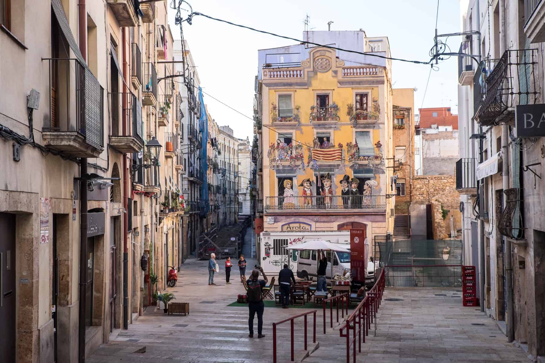 A man stands, taking a picture, in front of a building painted with a yellow art mural in Tarragona city, Spain. On either side are narrow avenues lined with old buildings.