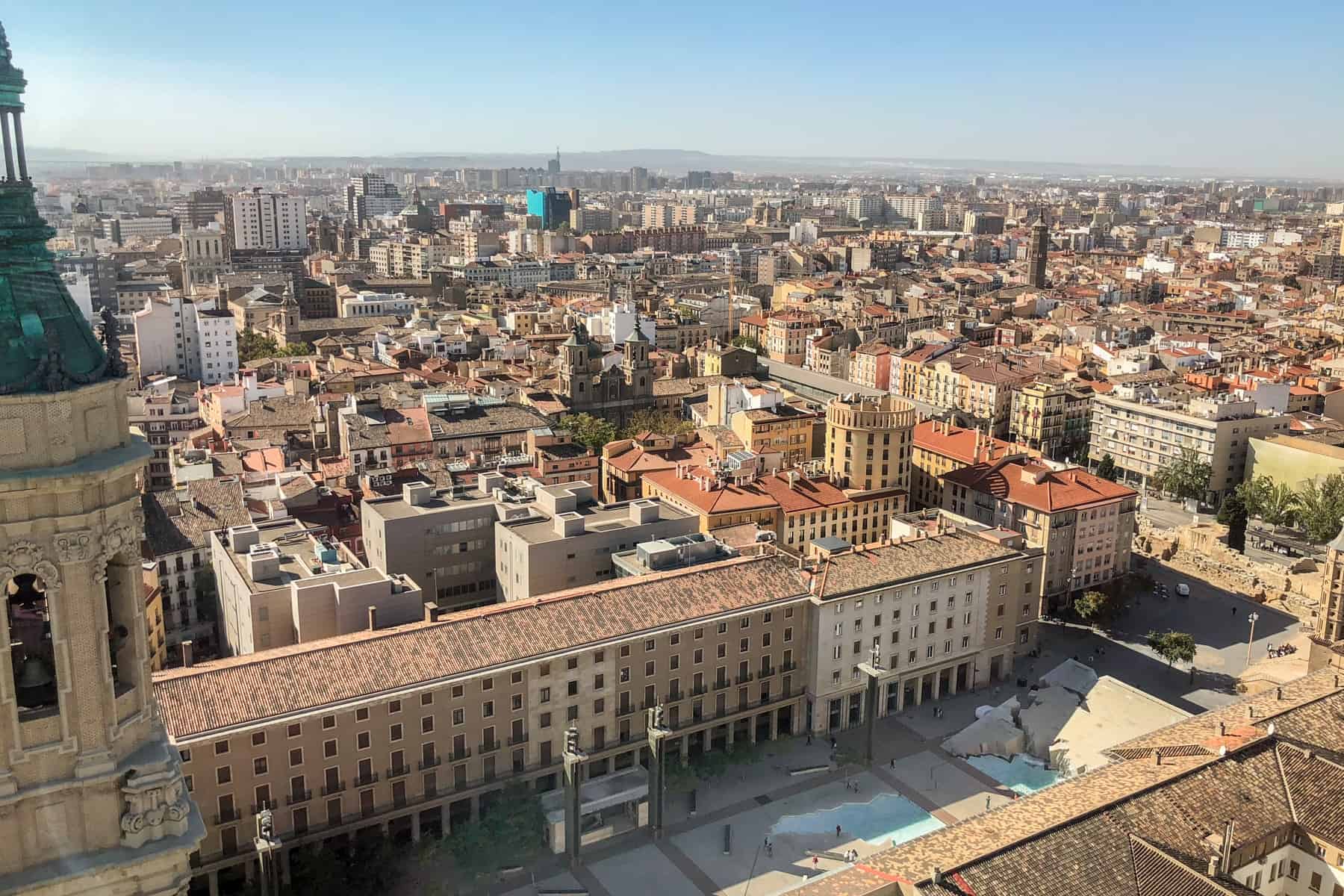 An elevated view of the densely packed Zaragoza city with a closer view down to the long building lined Plaza del Pilar square that features a blue Fuente de la Hispanidad water fountain in the shape of Latin America. 