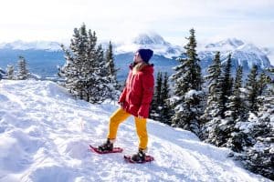 A woman in a purple hat, red jacket and yellow ski pants stands on a snowy hilltop backed by trees in snowshoes, in Banff National Park, Canada