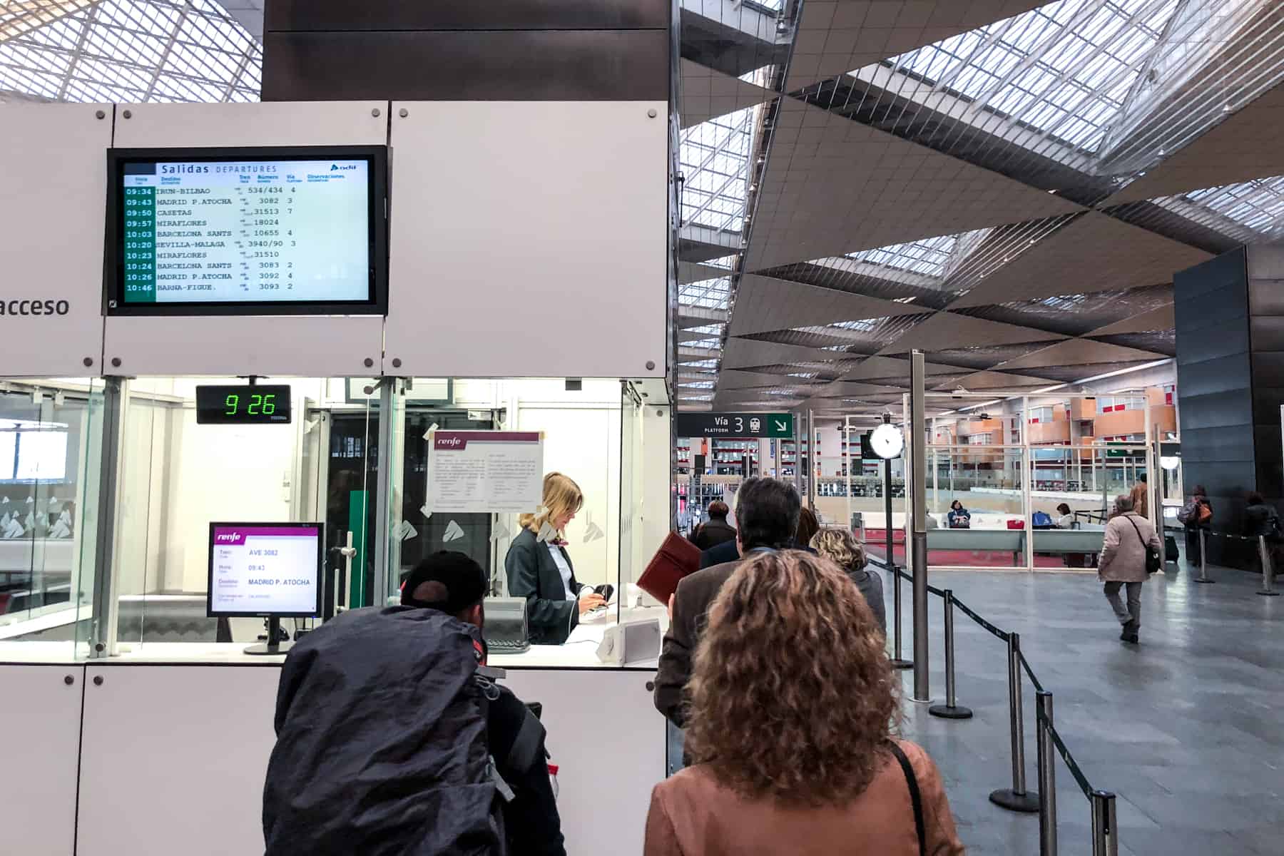 People queue up at a white ticket counter at a train station in Spain. 