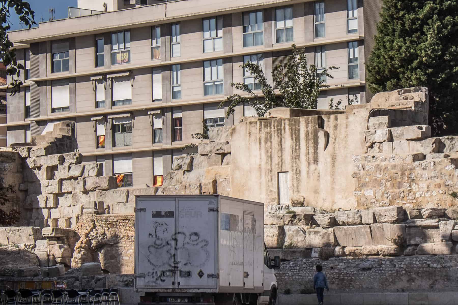 A large section of a Roman wall in Zaragoza city that stands in front of a modern apartment block. A white van is parked in front of the wall. 