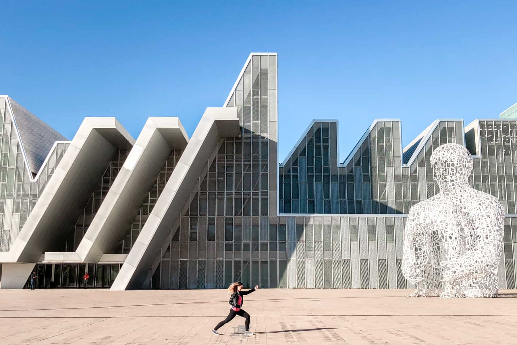 A woman wearing black and pink in a fighting stance pose in front of a wave-design building and a large figure made of white mesh metal. 