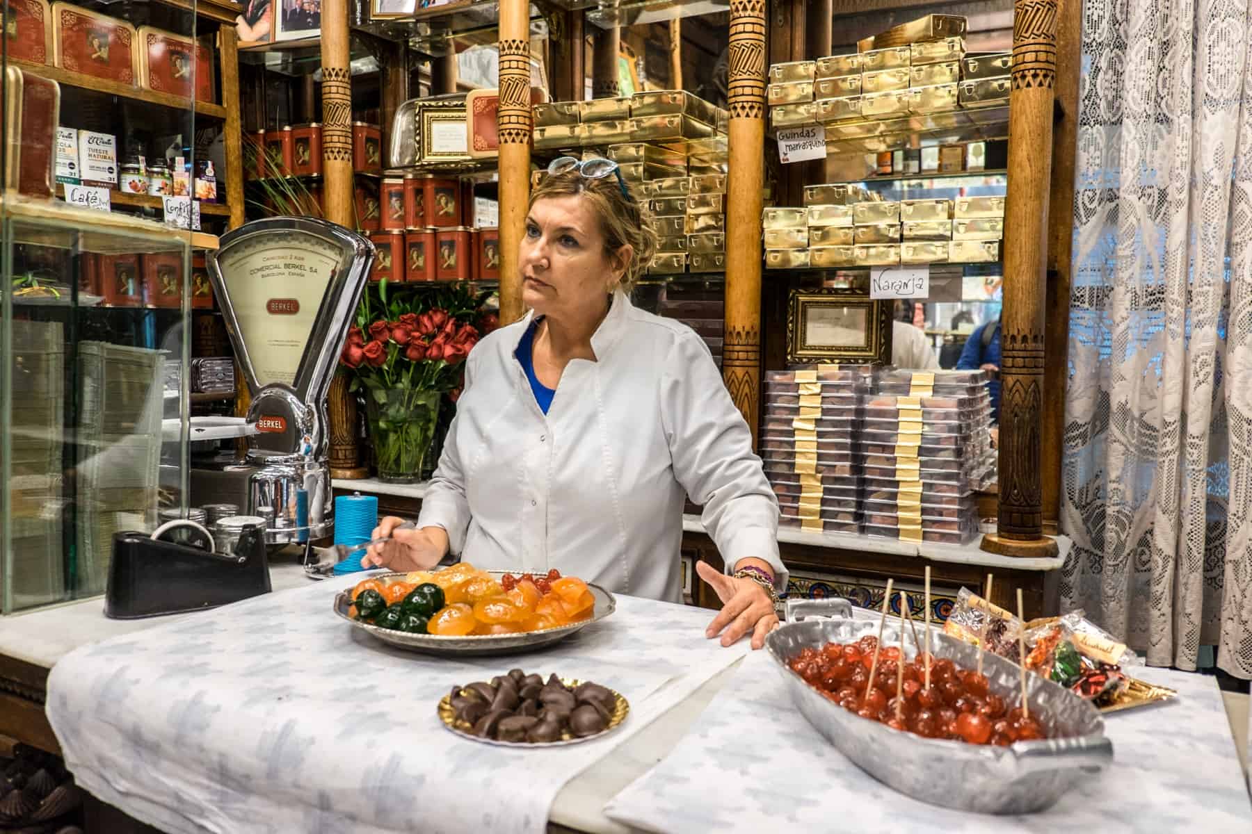 A woman in a white shirt stands shows off green, yellow and red candied sweets in a traditional confectionary store in Zaragoza. Behind her are carved wooden shelves full of boxes. 