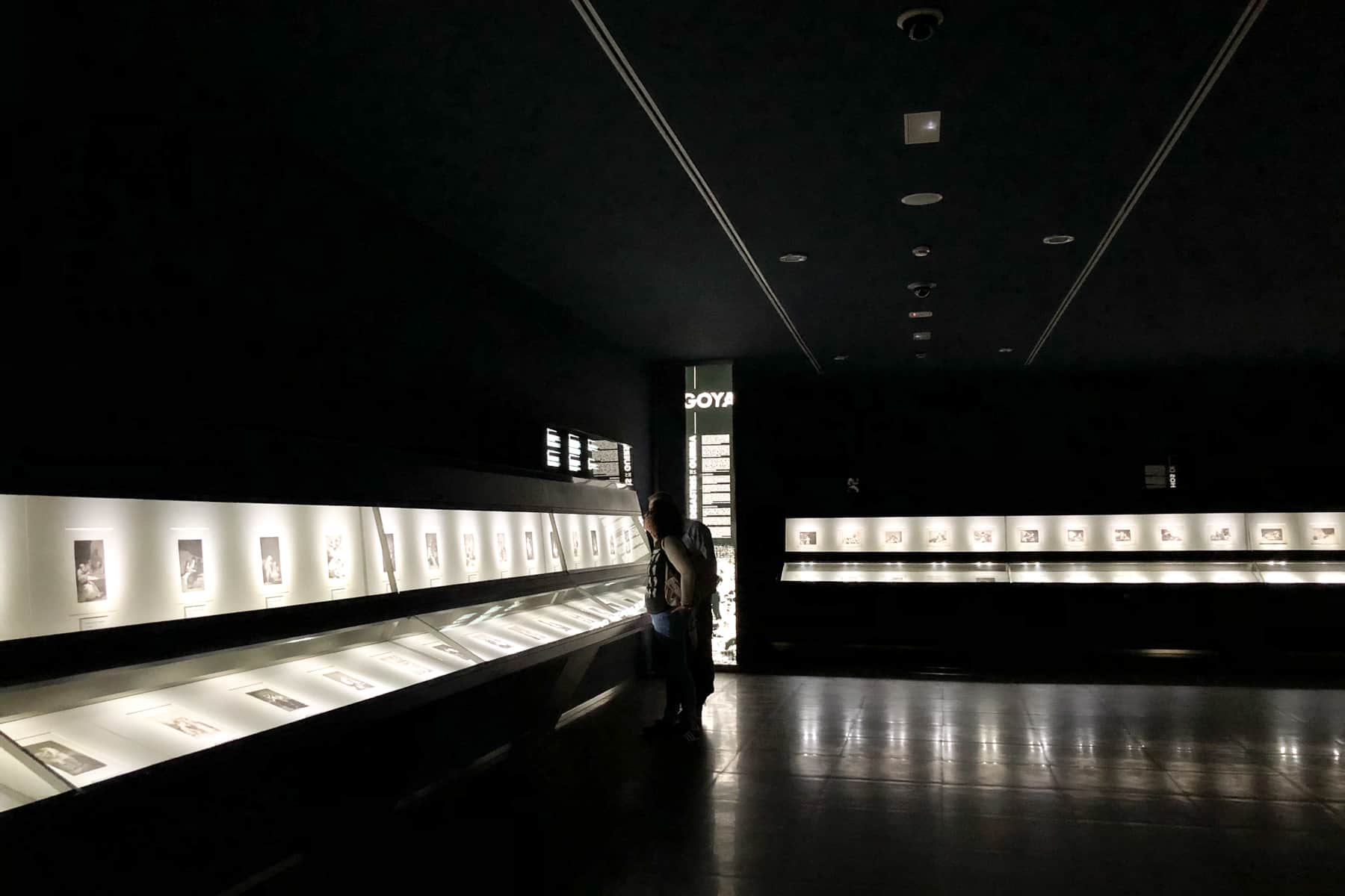 Two people in a black room looking at the long row of illuminated artworks of Goya, at the Goya Museum in Zaragoza. 