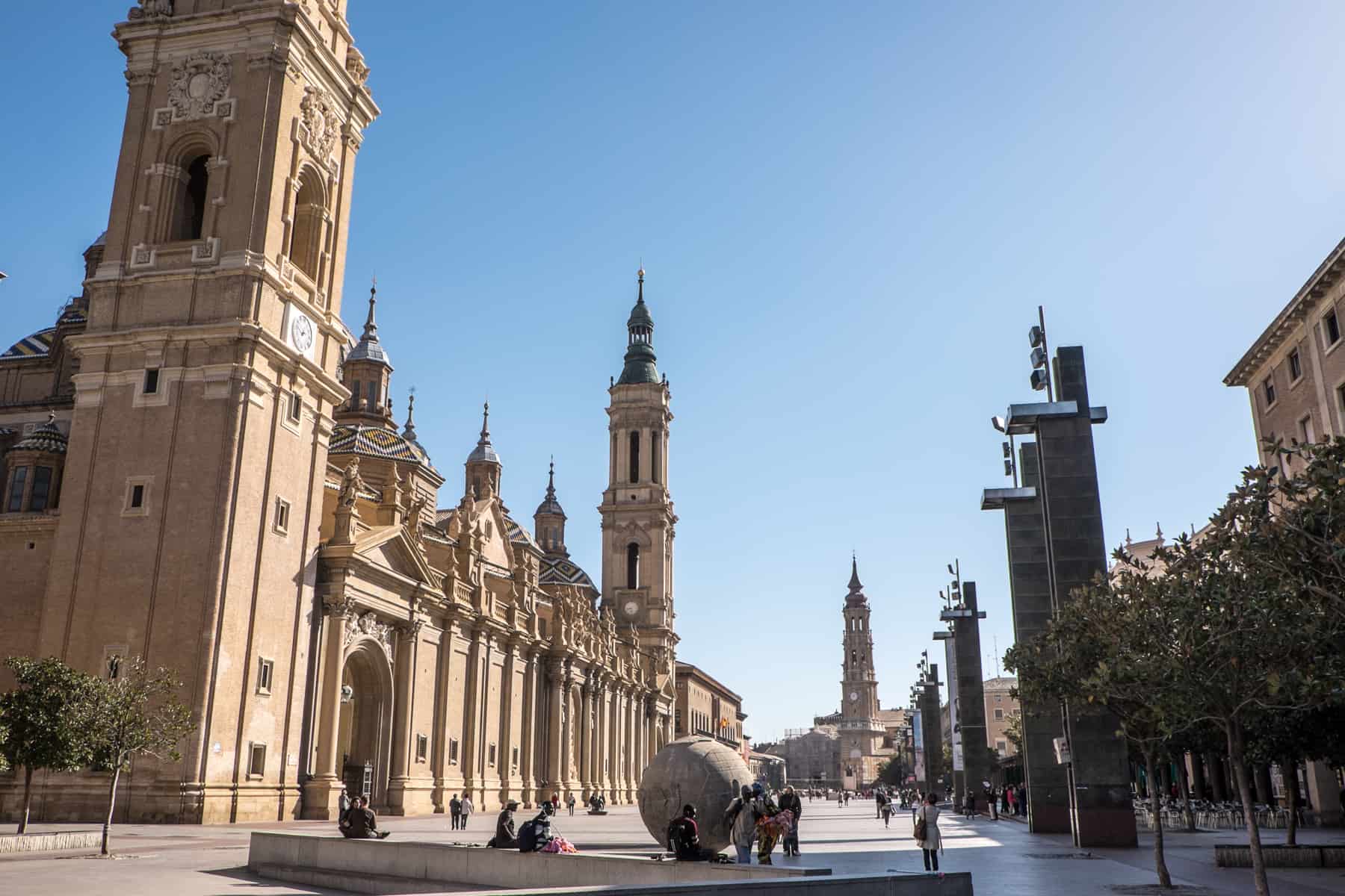 People gather around a globe artwork at one end of the long Plaza del Pilar square in Zaragoza, Spain. The dominant golden structure of The Cathedral-Basilica of Our Lady of the Pillar is on the left. 