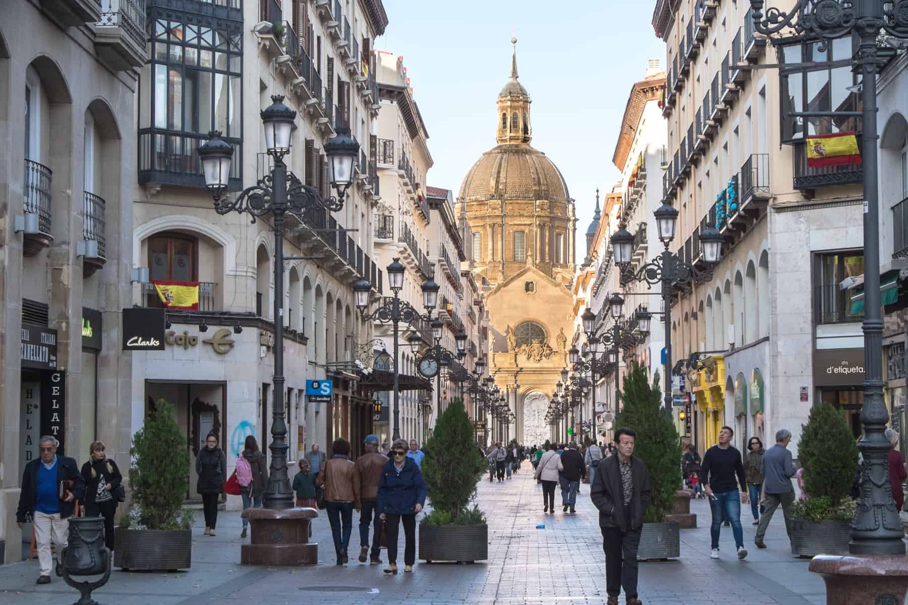 People walk down a street in Zaragoza city lined with white buildings and black lampposts. The street leads towards the golden dome structure. 