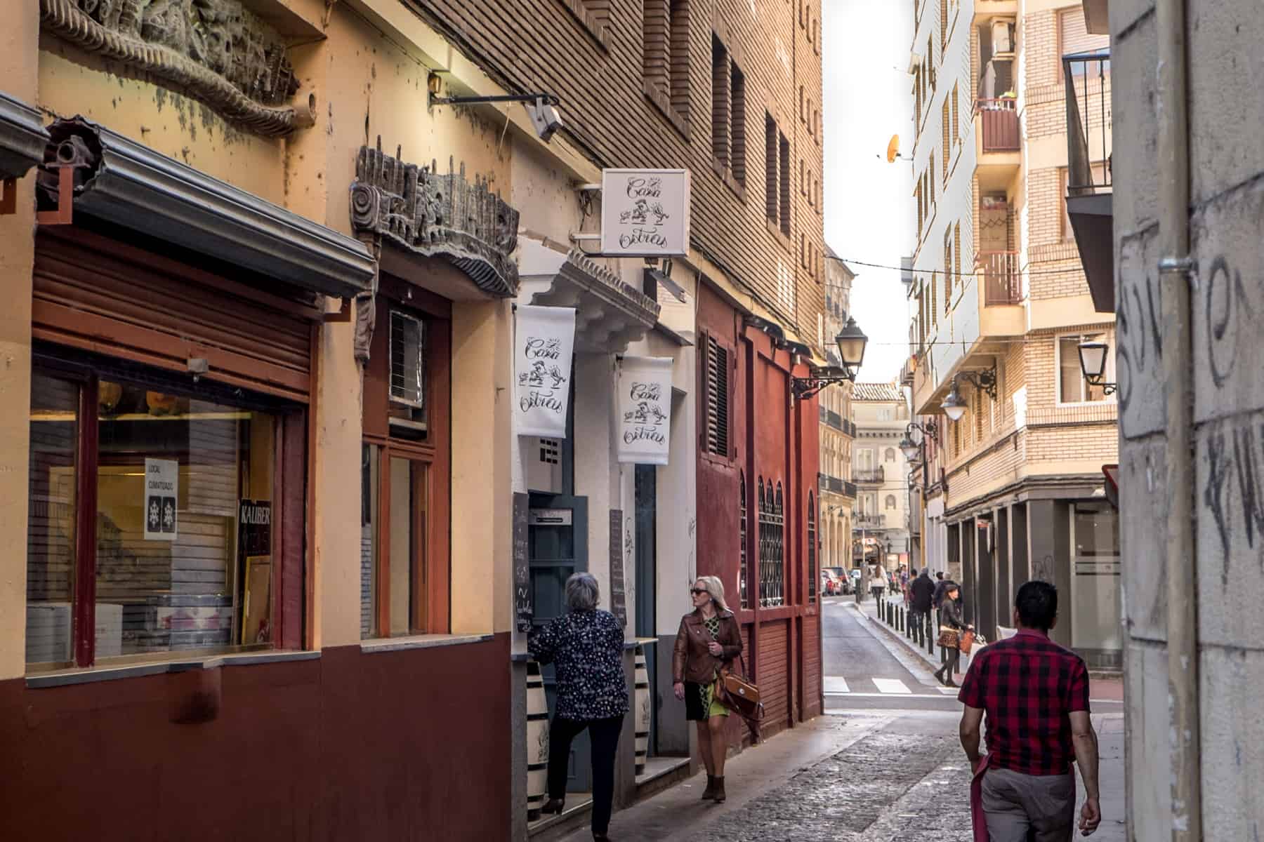 On a small side street in Zaragoza, two women stand outside a white building in between shop front painted in ochre red and yellow. A man walks past them. 