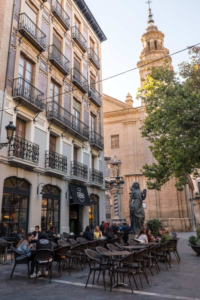 Outdoors seating at a cafe in front of a tall building with black balconies and next to a golden spired church. 