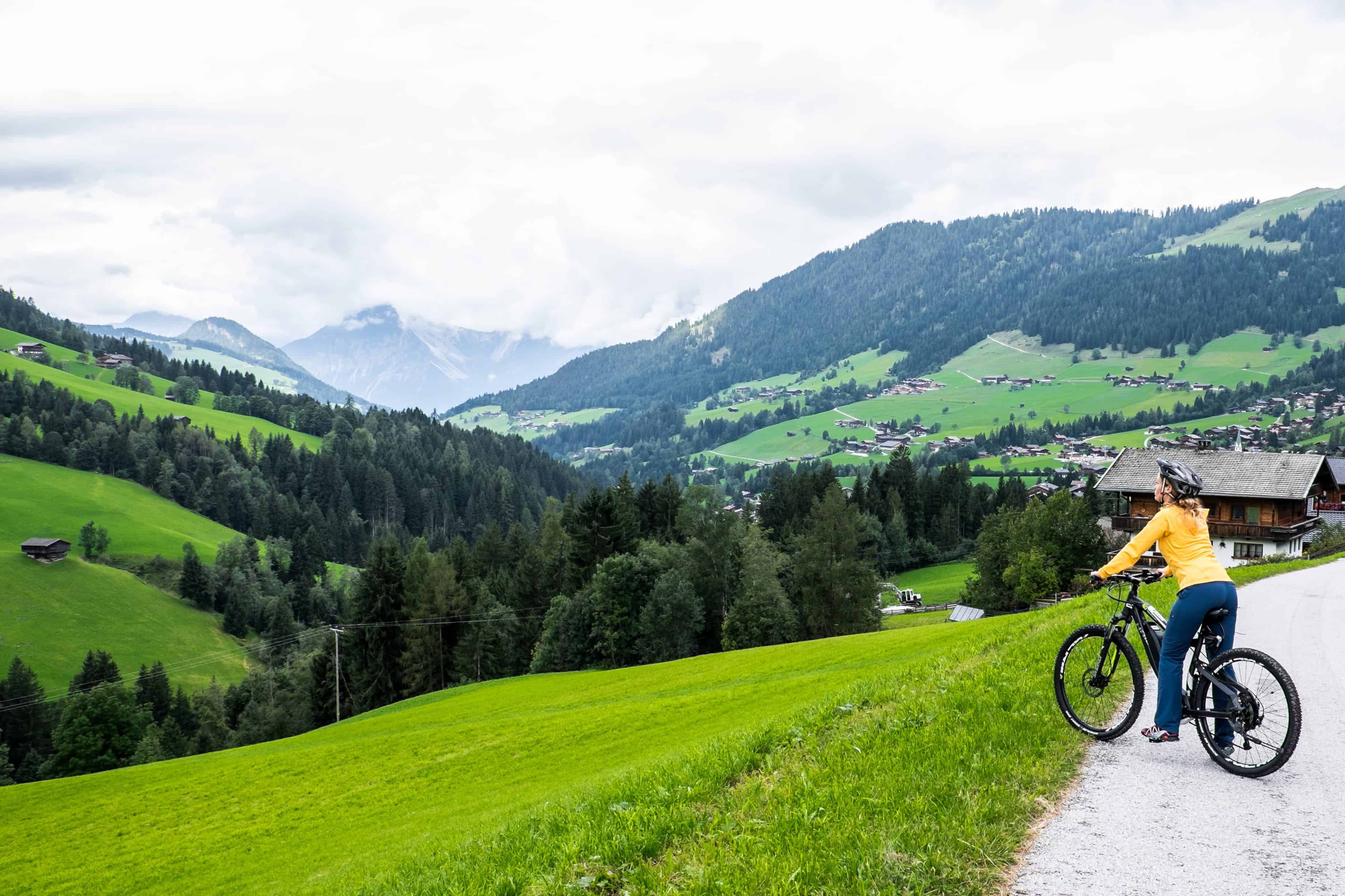 A woman with a yellow jacket and blue trousers on a mountain e-bike, stopping to look out over the mountain-backed green valley. 