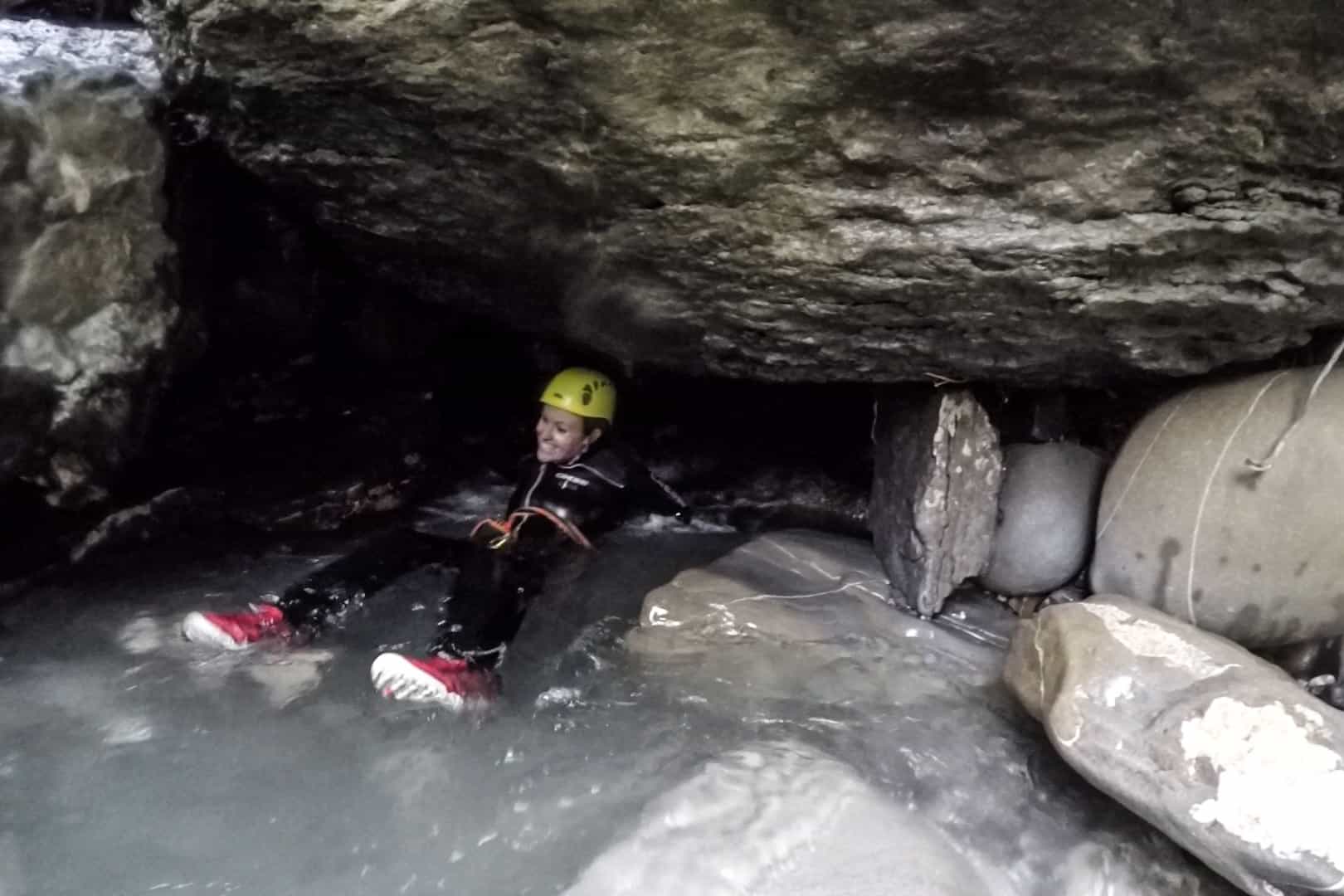 A woman in a black wetsuit, yellow helmet, and red shoes floats on her back through a narrow cave water passage.