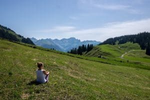 A women sits on a green hill looking out towards folds of mountains in the Bregenzerwald region, Vorarlberg, Austria