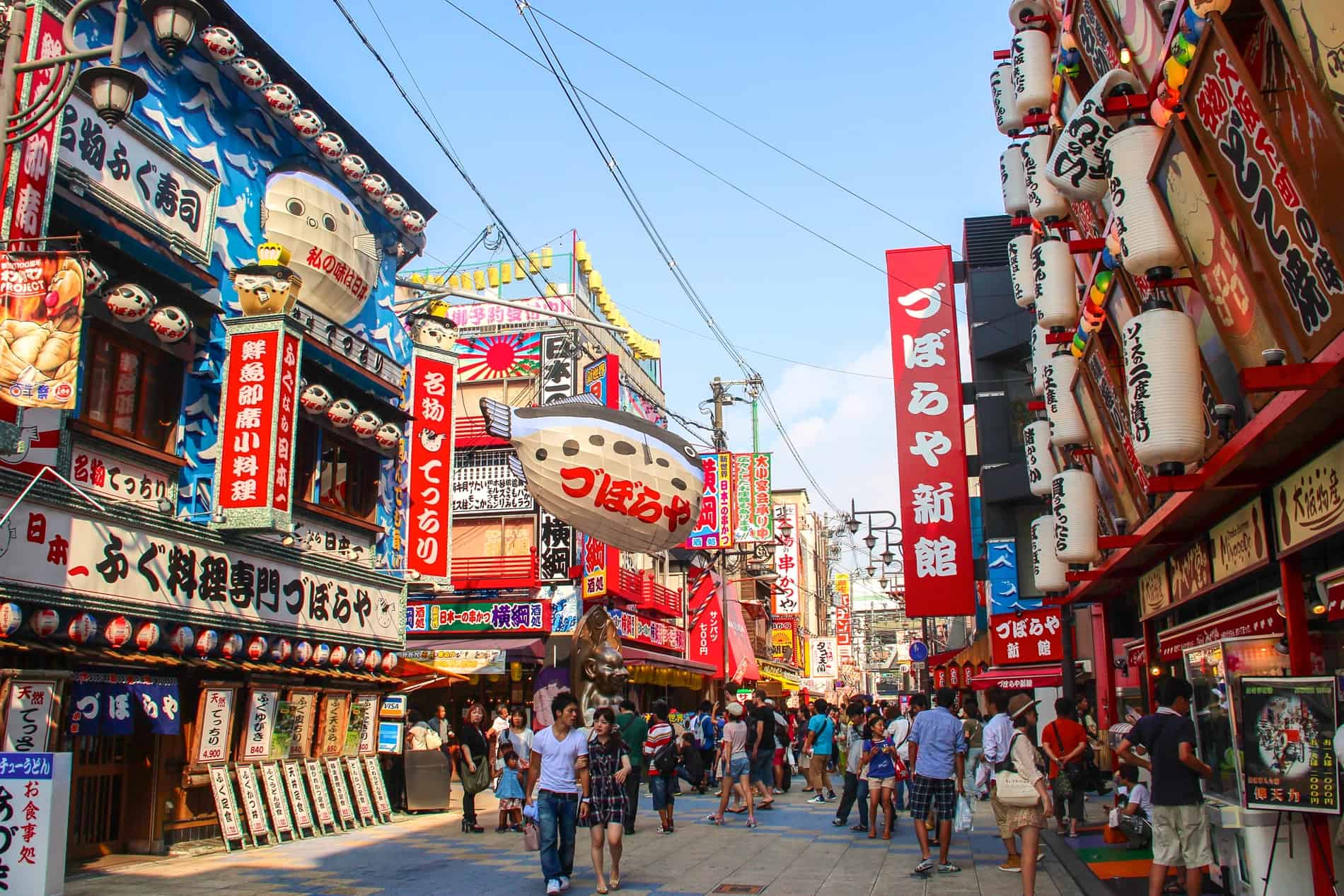 People walk through the famous Dotonburi street in Osaka during the day. Buildings are entirely covered in Japanese signs, and a giant white pufferfish hangs on a wire in the middle. 