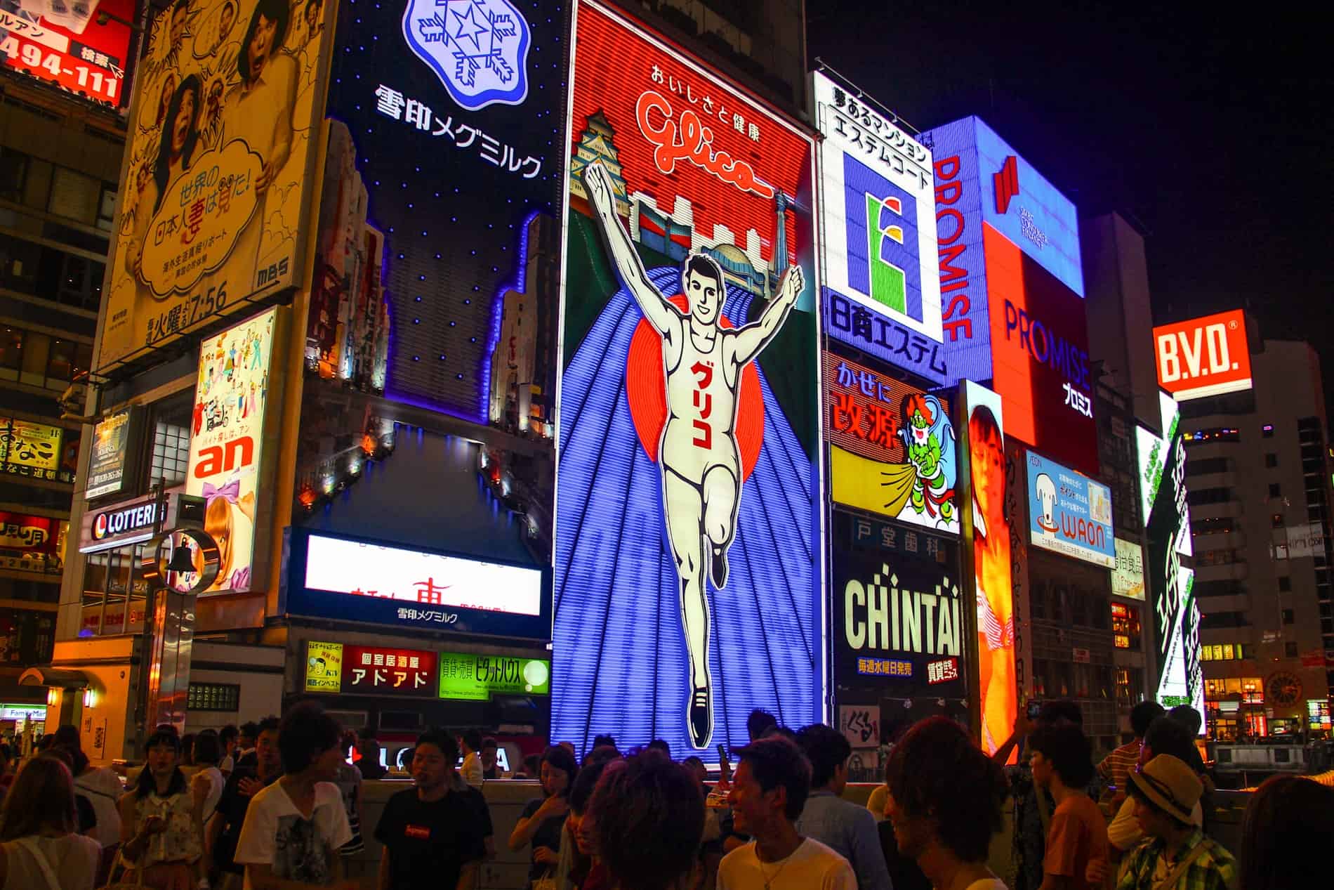 People gather in front of a building covered in animated advertising signs in Osaka, with the famous Glico Running Man sign in the middle.