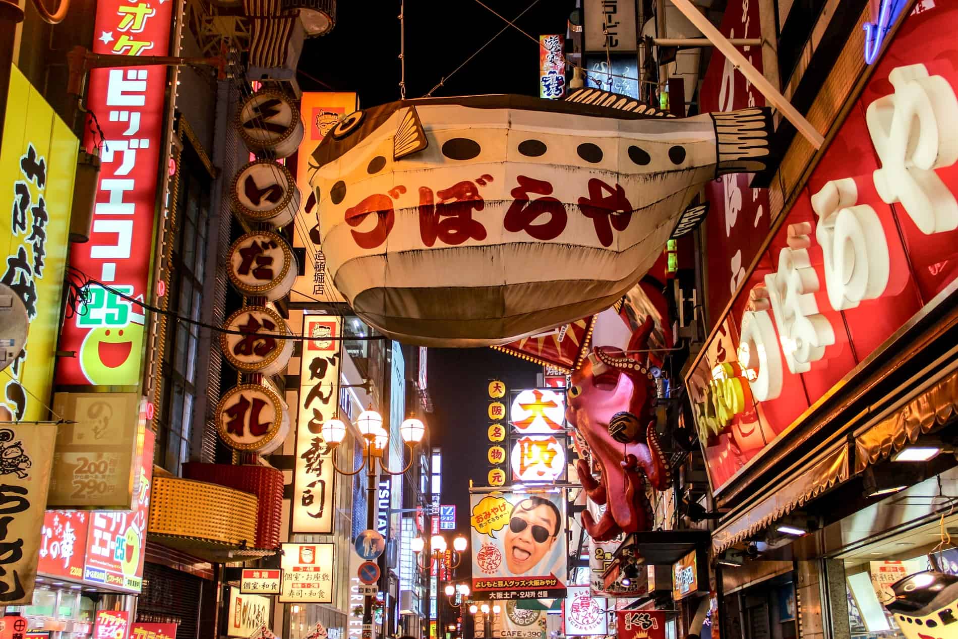 The Dotonburi area of Osaka at night. Buildings are entirely covered in lit-up signs, a giant white pufferfish hangs on a wire in the middle, and a giant pink octopus is stuck to a building on the right. 