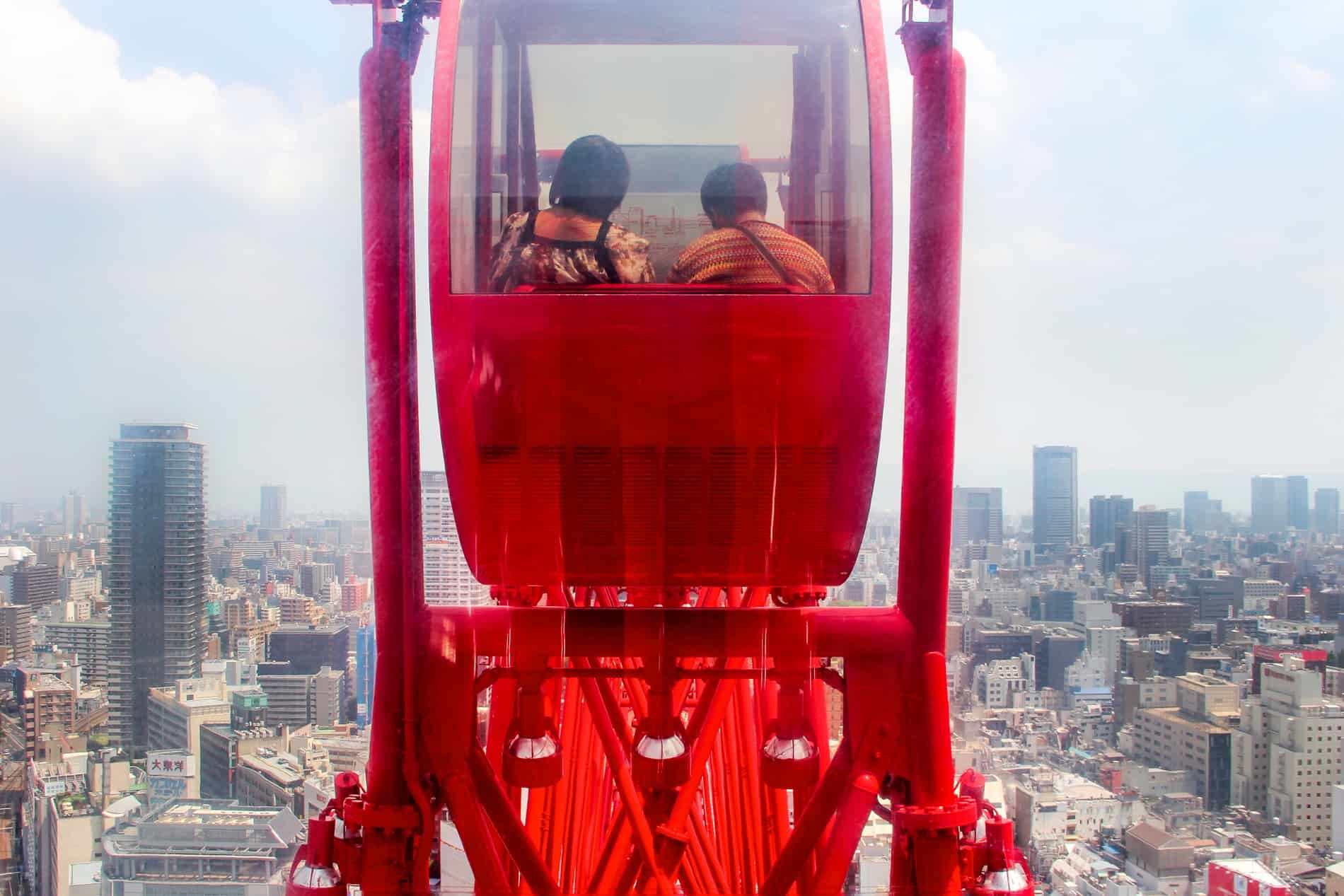 View into the red carriage of a Ferris Wheel carrying two passengers as it hovers above the city skyline of Osaka, Japan. 