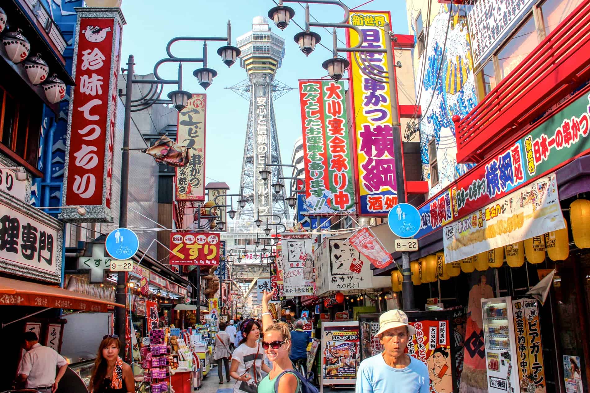A busy street in Osaka lined with chromatic signs in a multitude of a colours, with the silver Tsutenkaku Tower at the end. 