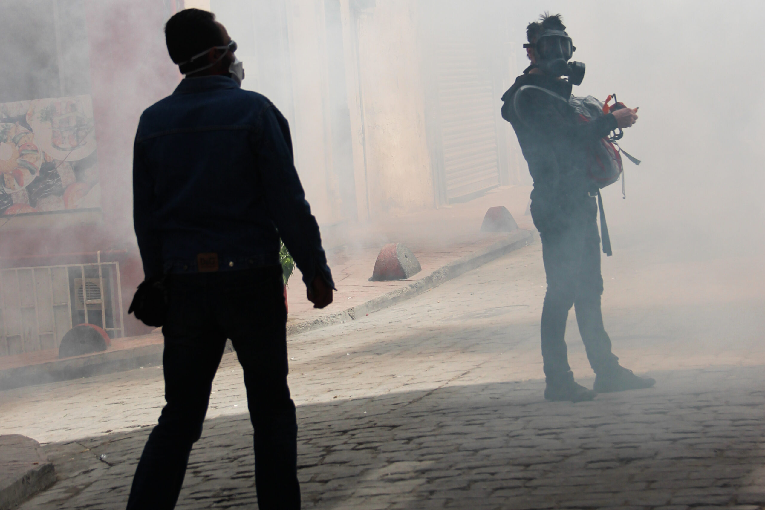 Rioters in dark clothing and wearing gas masks standing in a street filled with smoke.
