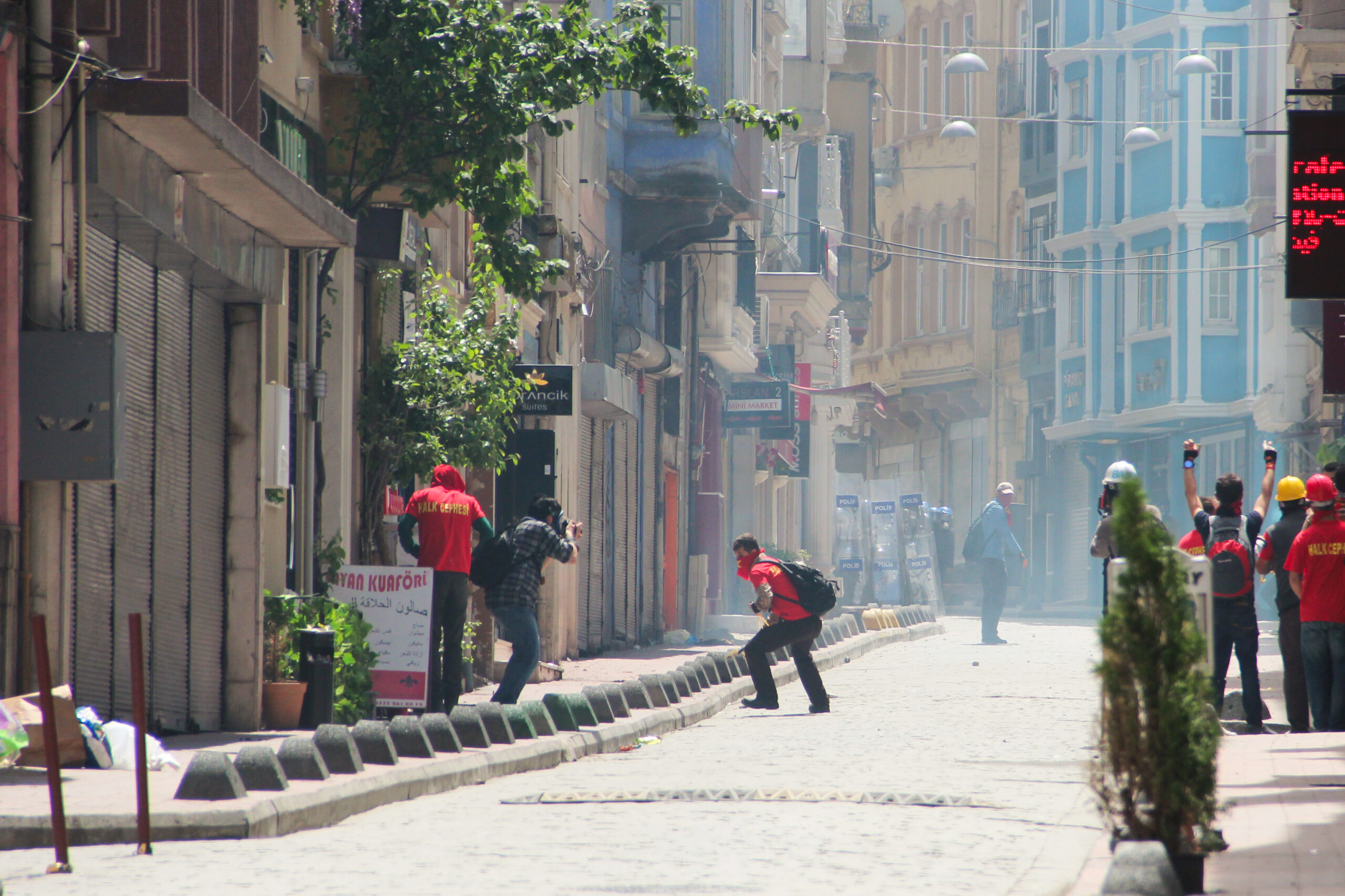 People in red shirts rioting in the Taksim neighbourhood of Istanbul as smoke and tear gas fills the air.