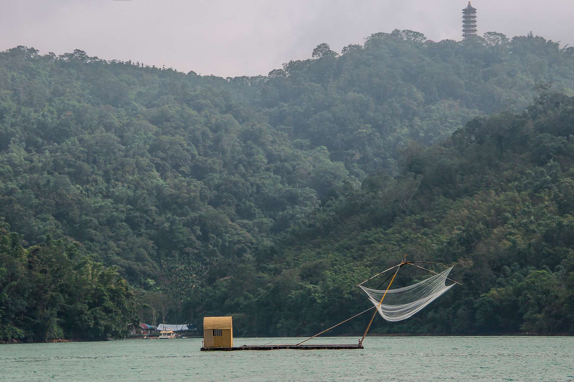 A fishing net in the middle on Sun Moon Lake in front of a large swathe of dense forest, topped with a Pagoda. 